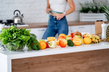 Diyet konsepti. Mutfakta sağlıklı yiyeceklerle güzel genç bir kadın var. Bel ölçümü yapıyorum. Meyveler ve sebzeler