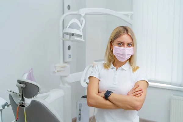 若い歯科医は現代の歯科クリニックで手を組んで立っています 顔のマスクをつけた医者 — ストック写真