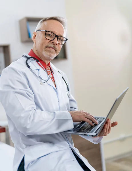 Zadowolony Dojrzały Lekarz Pracujący Laptopie Patrzący Kamerę Nowoczesnym Gabinecie Kliniki — Zdjęcie stockowe