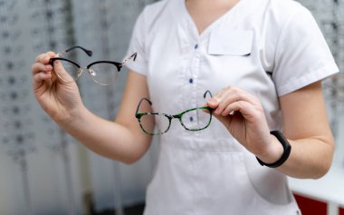 Woman shows two pairs of eyeglasses to choose from on a spectacles background in the optics. Close-up clipart