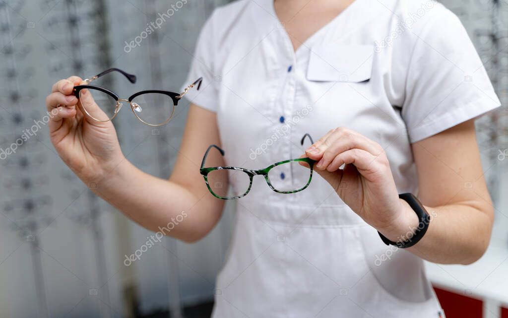 Woman shows two pairs of eyeglasses to choose from on a spectacles background in the optics. Close-up