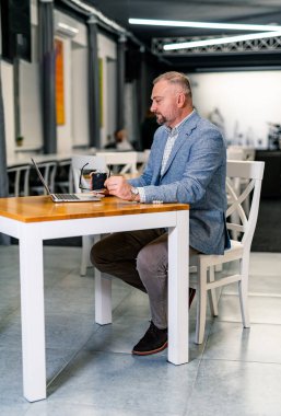 Kafedeki neşeli iş adamı laptopuyla çalışırken kahve içer. Takım elbiseli ve gözlüklü olgun bir erkek..