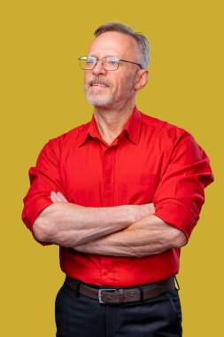 Kıdemli adam ellerini çapraz yapıp sarı arka planda duruyor. Kırmızı gömlek. Gözlüklü adam..