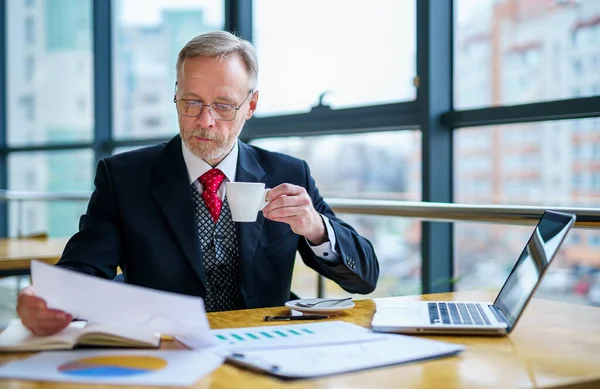 彼のオフィスで書類を見ている成熟した実業家 現代のオフィスでコーヒーを飲む男 パノラマシティビューの背景 — ストック写真
