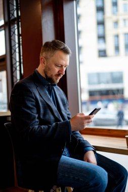 Genç işadamı bir kafede oturuyor ve telefonda mesaj yazıyor. Koyu renk ceketli ve kot pantolonlu yakışıklı adam..