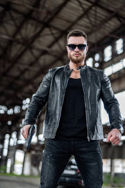 Handsome young man in leather jacket holding a gun. Cool sunglasses on brutal man. Brutal male in abandoned warehouse.