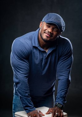 Sıradan kıyafetlerle poz veren Afrikalı Amerikalı kaslı adam. Siyah stüdyo geçmişi. Eller sandalyeye..