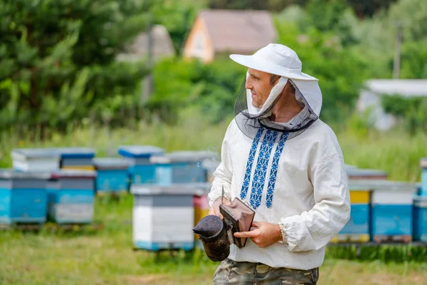 保護帽子の養蜂家は 手にミツバチのための喫煙者と立っています 背景にひづめ 夏の陽気な一日 — ストック写真