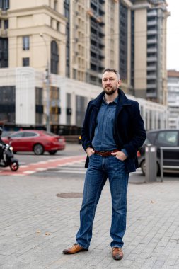 Gri ceketli ve kot pantolonlu bir iş adamı sokakta kameraya poz veriyor.