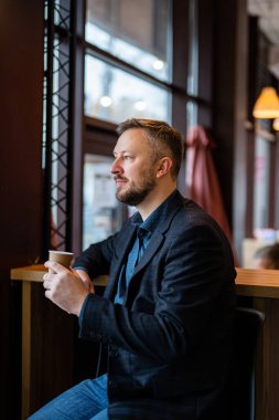 Yakışıklı sakallı erkek bir kafede bir fincan kahveyle oturuyor. Ceketli adam restoranın camına bakıyor.. 