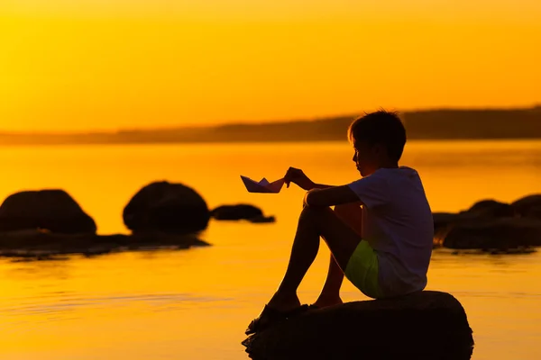 紙飛行機を手にした少年が川のそばの岩の上に腰を下ろしている 夏のオレンジの夕日 湖の近くの飛行機の折り紙と少年のシルエット — ストック写真