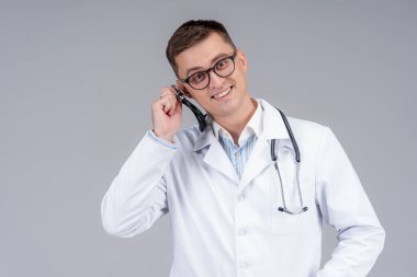 Yakışıklı, neşeli doktor kulağına fonendoskop dayadı. Beyaz önlüklü doktor açık gri arka planda poz veriyor.