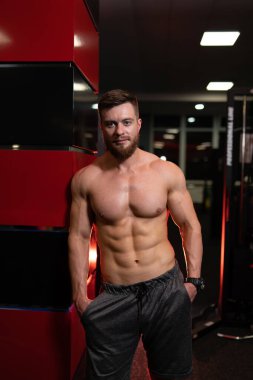 Sporcunun elleri cebinde spor salonunda kolona yaslanmış duruyor. Mükemmel vücudu ve güzel gülüşü olan gömleksiz yakışıklı bir adam..