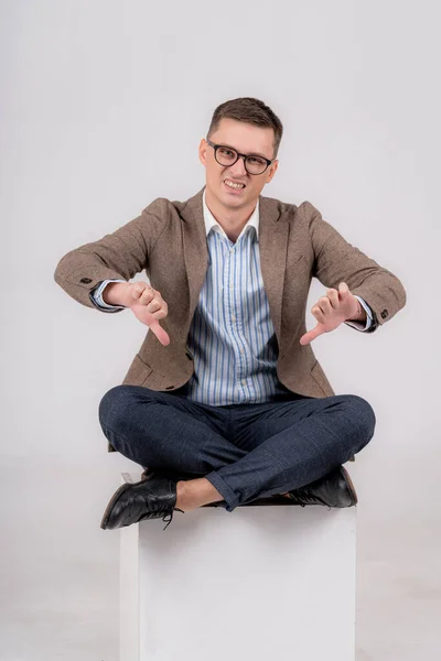 Formal man sitting on cube with legs crossed. Male showing both thumbs down