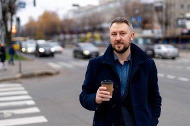 Kentin arka planında tek kullanımlık kağıt bardaktan kahve içen genç adam. Yaşam tarzı, içkiler ve insanlar konsepti.