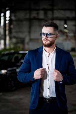 Mavi takım elbiseli moda adamı hasarlı deponun içinde poz veriyor. Erkek, beyaz gömlek ve modern gözlük takıyor. Siyah arabalı bulanık arkaplan.