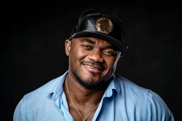 Man in fashion leather cap and blue shirt poses to camera. Head and shoulder portrait of african american on black background.