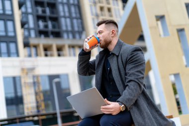 Takım elbiseli genç profesyonel iş adamı dışarıda yüksek teknolojili laptop kullanıyor. Bir mola sırasında dışarıda çalışan başarılı bir yönetici ve dizüstü bilgisayarda internette bilgi arıyor.