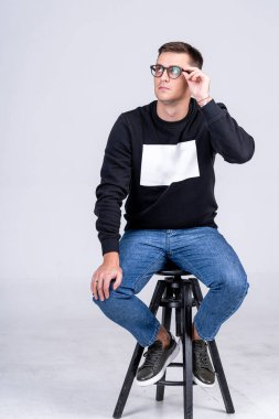 Genç ve çekici beyaz adam modern kapüşonlu yüksek bar sandalyesinde oturuyor ve kameraya poz veriyor. Beyaz arka plan. Yakın çekim..