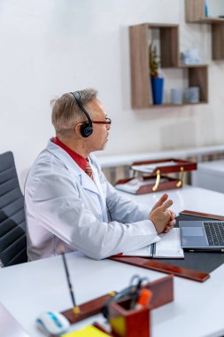 Doktor dizüstü bilgisayarla muayene yapıyor. Önlüklü deneyimli doktor kulaklıklarla masada oturuyor. Yakın çekim..