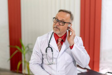 Olgun doktor ofisinde oturuyor. Boynunda steteskop olan beyaz önlüklü ve gözlüklü bir doktor. Yakın çekim..