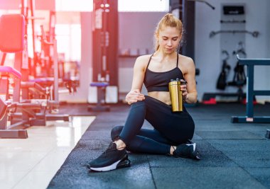Kadın antrenmandan sonra yerde oturur ve spor bardağından su içer. Modern spor salonundaki seksi zayıf fitness kızı. Yakın çekim..