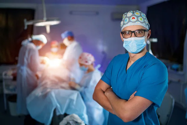 Portrait of doctor wearing eye glasses in blue uniform standing cross hands in operation room. Concept of doctors, patients, medicine and operating theater.
