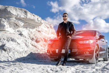 Modern arabanın yanında duran yakışıklı adam. Beyaz dağların nefes kesici manzarası. Sıkıntılı kot ceketli ve güneş gözlüklü adam..
