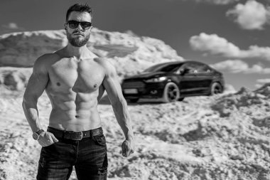 Kaslı vücut yapısı olan güçlü bir adam. Günümüz arabasının önünde duran güneş gözlüklü vücutçunun tek renkli fotoğrafı. Güzel bir yer, beyaz dağlar ve gökyüzü..