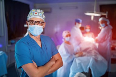 Doktor maske takarak el ele tutuşuyor. Arka planda tıbbi ekip var. Yüzünde tıbbi maske olan gözlüklü bir doktor. Yakın çekim.