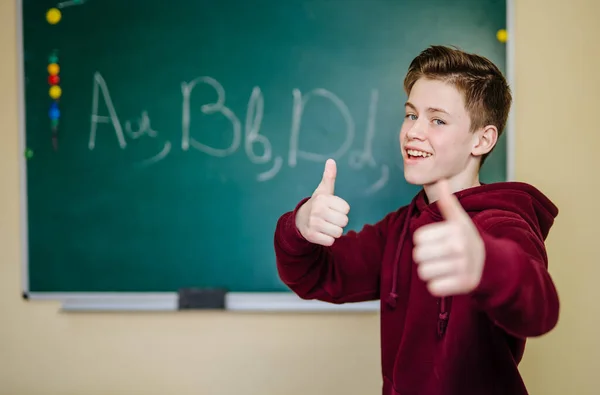 Student of higher school is standing in dark hoodie near the green blackboard in the classroom and showing big thumbs. School education concept