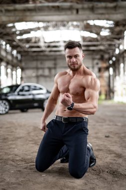 Lüks arabanın yanında dizlerinin üstünde duran havalı üstsüz adam. Atletik vücutlu yakışıklı bir adam kameraya poz veriyor.