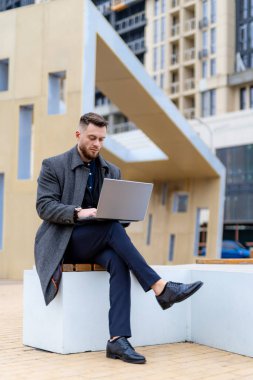 Dışarıda dizüstü bilgisayarda çalışan yakışıklı bir gencin portresi. Moda adamı şehir merkezinde bankta oturuyor. Bulanık arkaplan