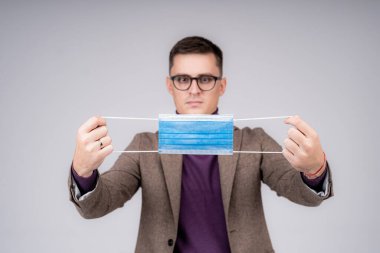 Yakışıklı iş adamı koruyucu maske tutuyor ve açık gri arka planda izole edilmiş bir şekilde ona bakıyor. Takım elbiseli genç adam stüdyoda duruyor.