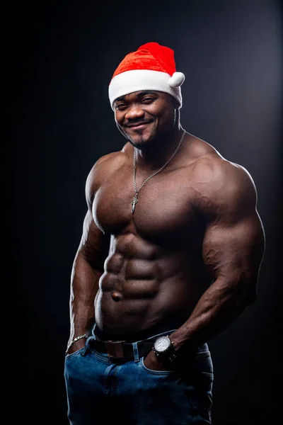 一个身强力壮的非裔美国人 戴着圣诞帽 背对着背景展示他的体形的画像 男人完美的身体在工作室摆姿势 同时看着相机 — 图库照片
