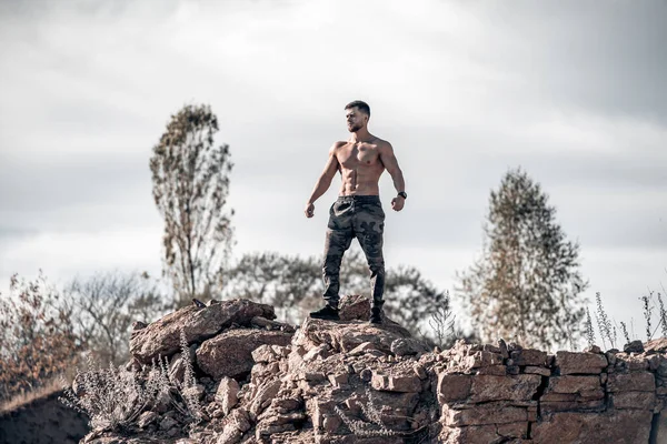 Strong bearded man in military pants. Bodybuilder standing on a rock and looking away. Naked torso. Full length view