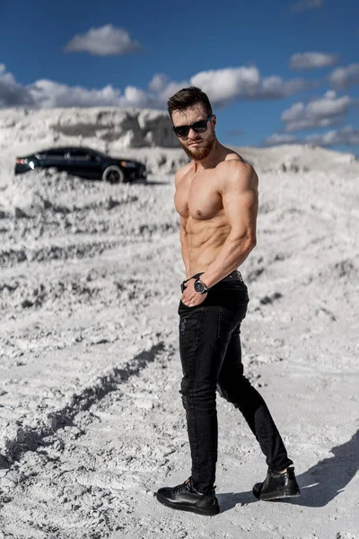 Brutal man with no shirt is standing on the road near his car. Man in front of the luxury auto. Horizontal photo. Full length view