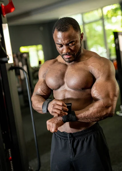 Vertical view of the young multiracial man with naked torso putting special bandages at his hands on modern gym background. Strength and motivation concept