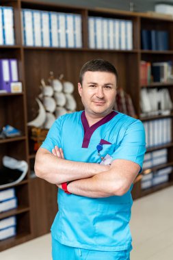 Hastane koridorunda kameraya bakıp gülümseyen erkek cerrahın portresi. Modern hastanede mavi elbiseli doktor kollarını kavuşturdu. Tıp konsepti