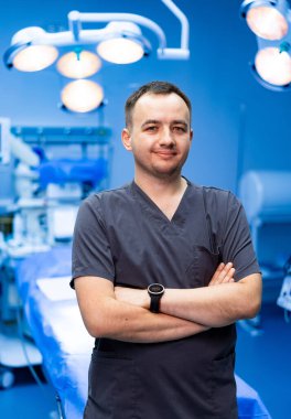 Ameliyathanede kendinden emin bir erkek doktor var. Kolları kavuşturulmuş, kendine güvenen bir erkek doktor ameliyat ışıkları altında modern bir ameliyathanede duruyor..