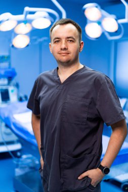 Ameliyathanede poz veren erkek doktor. Koyu renk önlüklü bir erkek doktor modern bir ameliyathanede rahatça poz veriyor..