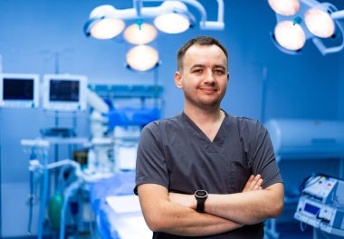 Cerrahi odasında ekipmanlı bir erkek doktor. Ameliyathanede kollarını kavuşturmuş bir erkek doktor cerrahi ekipmanların önünde duruyor..