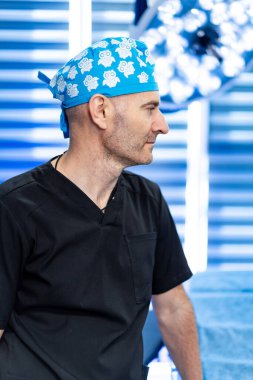 Mavi cerrahi şapkalı siyah önlüklü bir cerrah. Siyah önlüklü ve mavi desenli cerrahi şapkalı erkek cerrah modern ameliyathanede duruyor..