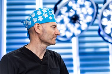 Hastanedeki mavi cerrahi şapkalı cerrahın portresi. Siyah önlüklü, arka planda ışıklar olan mavi cerrahi şapkalı cerrahın yakın çekimi..