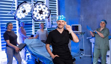 Cerrah ameliyathanede ekiple poz veriyor. Cerrahi şapkalı ve siyah önlüklü bir doktor modern cerrahi odasında tıbbi ekiple poz veriyor..