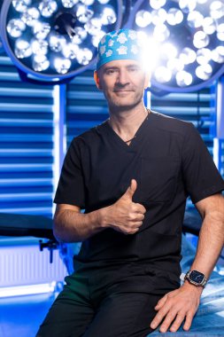 Cerrah ameliyathanede onay veriyor. Siyah önlüklü bir cerrah ameliyathanede arka planda cerrahi ışıklar varken onay veriyor..