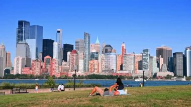 New Jersey, ABD, 19 Ağustos 2025: İnsanlar yürür, oturur ve Jersey City, ABD 'deki rıhtımda fotoğraf çekerler. Güneşli bir günde nehrin yanında sakin bir gün. Arkaplanda ikonik Manhattan silueti.