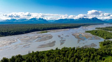 Alaska 'nın havadan görünüşü nehir ve dağ sıralarını örüyor. Alaska nehri, arka planda kum yığınları, yoğun yeşil orman ve dağ sıralarıyla örüldü..