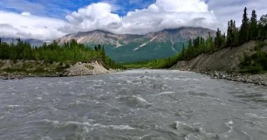 Kayalık kıyılar arasında çalkantılı bir nehir. Pofuduk bulutlar arka plandaki muhteşem dağların tepelerini kaplıyor. Alaska, ABD.