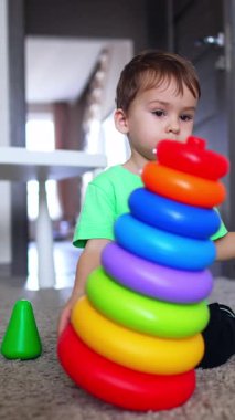 Oyuncak piramidi yere iten ve parçalara ayıran güzel çocuk. Beyaz çocuk evde oynuyordu. Dikey video